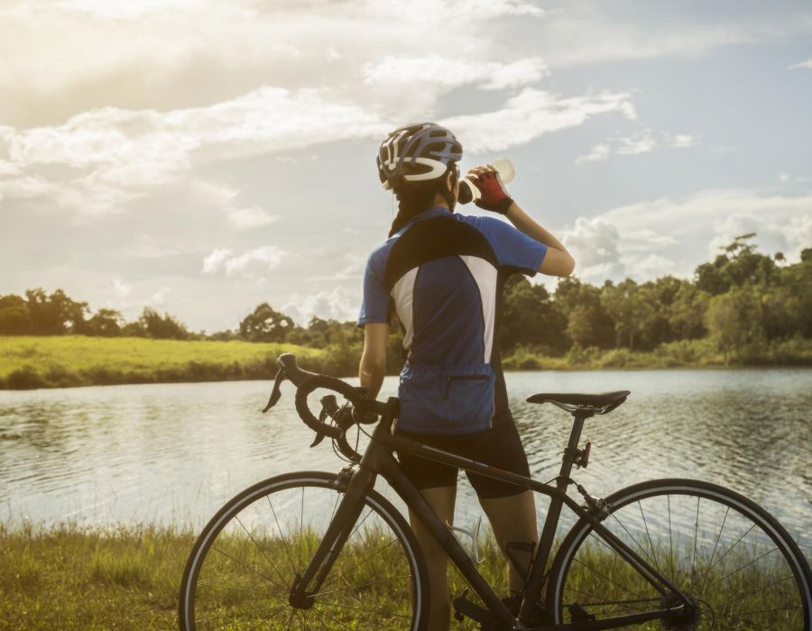 Woman cyclist standing next to her bicycle while taking a drink from a water bottle and looking out at a river view